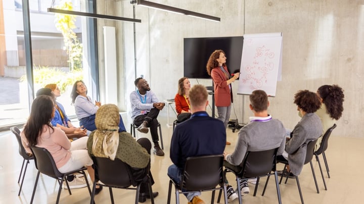 A diverse group of people seated in a semi-circle during a workplace training session, while a woman stands at a flip chart presenting and pointing to diagrams.