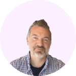 The image shows a middle-aged man with short gray hair and a beard, wearing a checkered shirt and smiling at the camera.