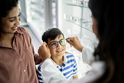 A youg boy being fitted for glasses in a shop. His mother stands beside him.