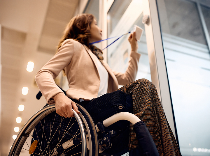 A person in a wheelchair reaching up to open a door with a keycard, wearing a beige coat and looking out the window.