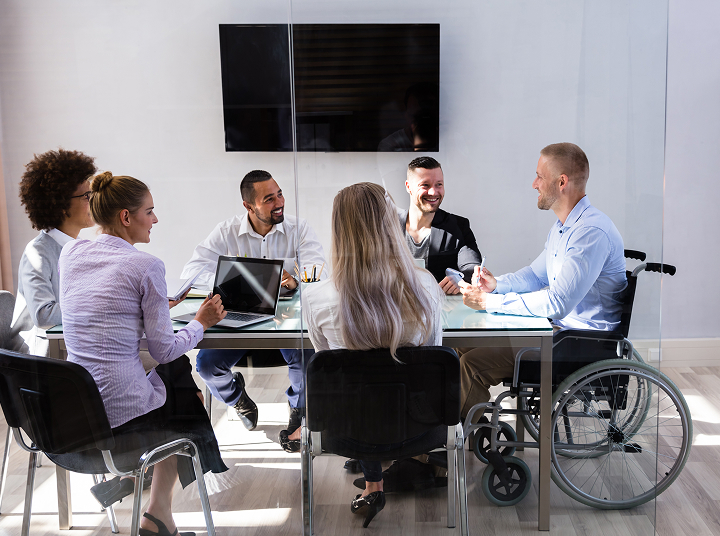 A group of diverse professionals engaged in a meeting around a glass conference table in a modern office. One man in a wheelchair is actively participating in the discussion, while others listen, smile, and use laptops or notebooks. A large blank screen is mounted on the wall behind them.