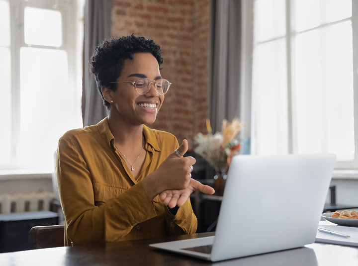 A smiling person with short curly hair and glasses sitting at a desk, working on a laptop computer in a cozy office setting.