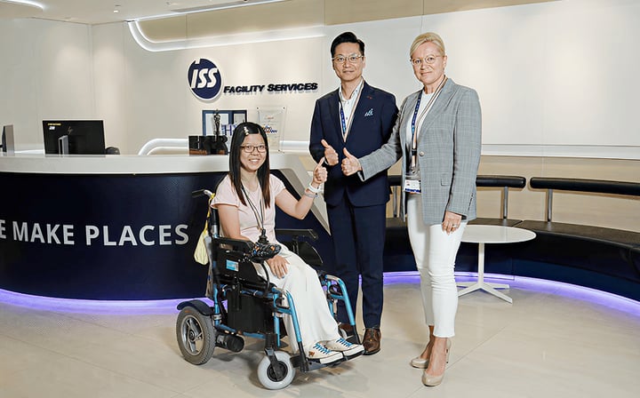 The image shows three people standing in an office setting. One person is seated in a wheelchair, while the other two are standing next to them. The office has a sign for "ISS Facility Services" visible in the background.