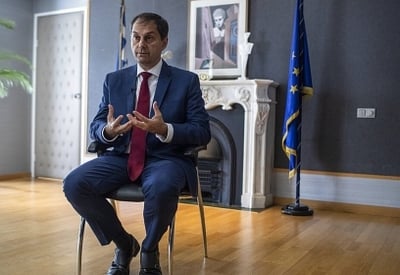 A man in a suit and tie sitting in a chair in an office, with a European flag visible in the background.