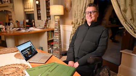 The image shows a man - Mobility Mojo Co Founder and CEO Stephen Cluskey, sitting in a cozy home office, wearing glasses and a black sweater. He is seated in a chair and appears to be working on a laptop computer on a desk in front of him. The room has a warm, inviting atmosphere with bookshelves and curtains visible in the background.