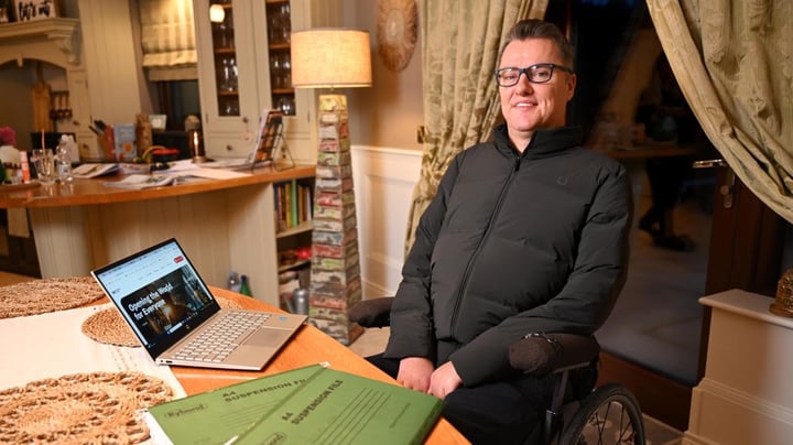 The image shows a man - Mobility Mojo Co Founder and CEO Stephen Cluskey, sitting in a cozy home office, wearing glasses and a black sweater. He is seated in a chair and appears to be working on a laptop computer on a desk in front of him. The room has a warm, inviting atmosphere with bookshelves and curtains visible in the background.