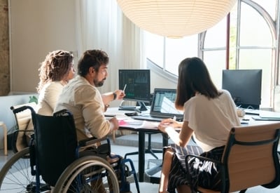 The image shows a group of people working together in an office setting. There are several people seated at desks, including one person in a wheelchair, and they appear to be collaborating on computer screens and documents.