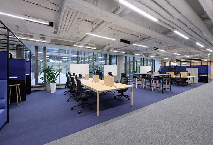 This image shows a modern, open-plan office space with a variety of workstations, desks, and seating areas. The space has a clean, minimalist design with exposed ceilings, large windows, and a blue and gray color scheme.