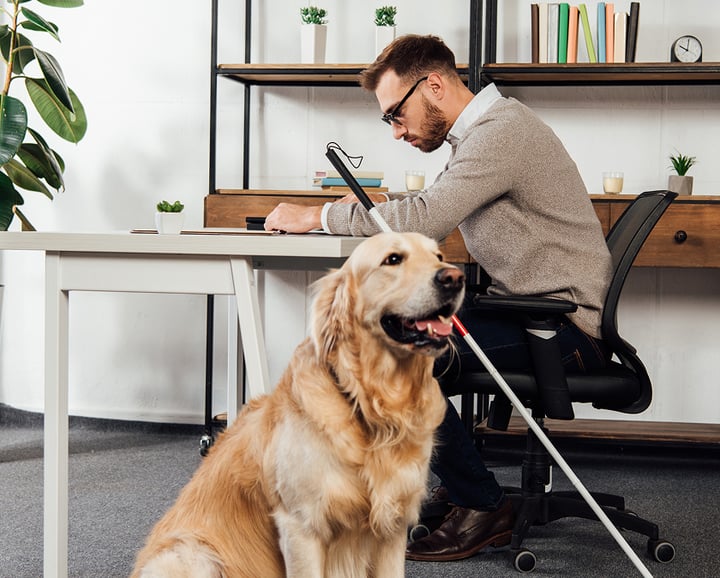 A man in a gray sweater is working at a desk, with a golden retriever sitting next to him. The dog appears to be a service animal, providing companionship and assistance to the man.
