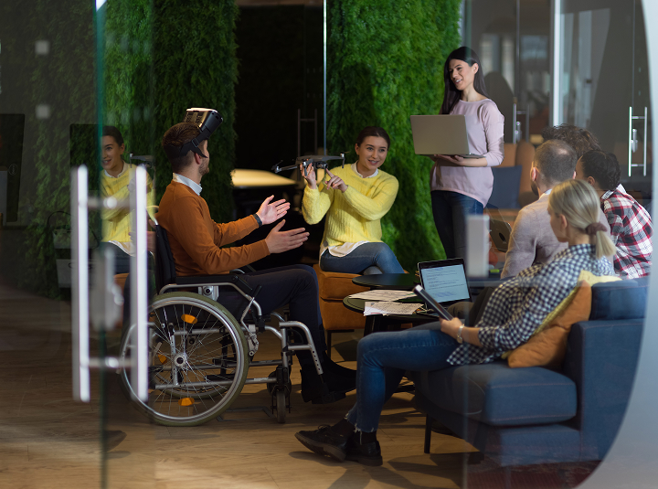A diverse group of people in a modern office setting engaged in a tech-focused meeting. A man in a wheelchair is wearing a headset, while a woman next to him holds a drone and smiles. Others are seated around, using laptops and listening to a standing woman who is presenting with a laptop in hand. Green plant walls create a natural backdrop.