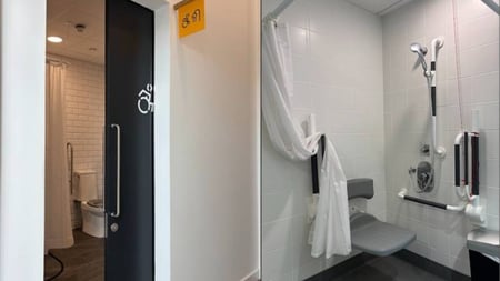 Accessible bathroom with white tile, wall-mounted shower fixtures, fold-down gray shower seat, and grab bars. A black door with accessibility symbol opens to a separate toilet room with wood walls.