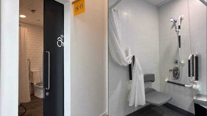 Accessible bathroom with white tile, wall-mounted shower fixtures, fold-down gray shower seat, and grab bars. A black door with accessibility symbol opens to a separate toilet room with wood walls.