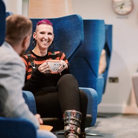 A woman sitting in a chair holding a mug with a big smile on her face sitting opposite someone who is turned away from the camera