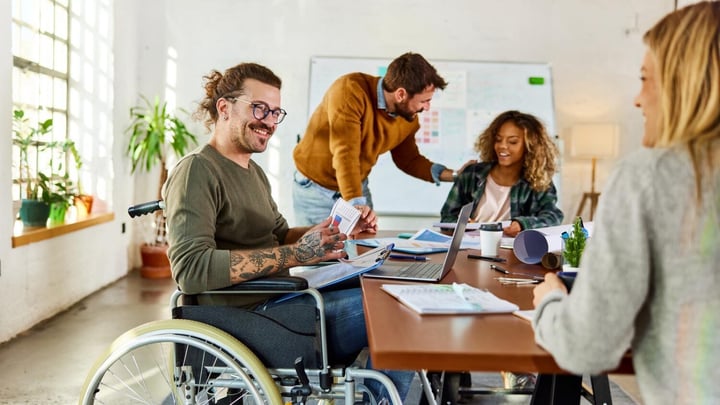 A diverse group of colleagues in a bright, modern office having a collaborative meeting. A man in a wheelchair is smiling and holding documents while seated at the table with a laptop. Others are engaged in discussion and working with papers and charts.