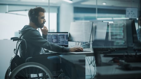 A man with a beard wearing a grey hoodie and large over-ear headphones sits in a wheelchair at a modern office desk. He is focused on several computer monitors displaying lines of code. The background shows a bright, professional open-plan office setting with glass partitions.