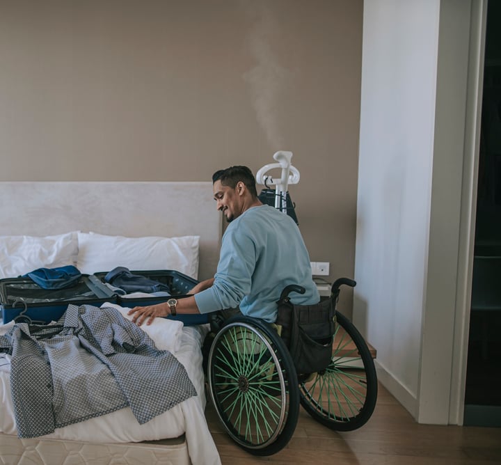 A person in a wheelchair using a laptop in a hotel bedroom with a lowered bed.