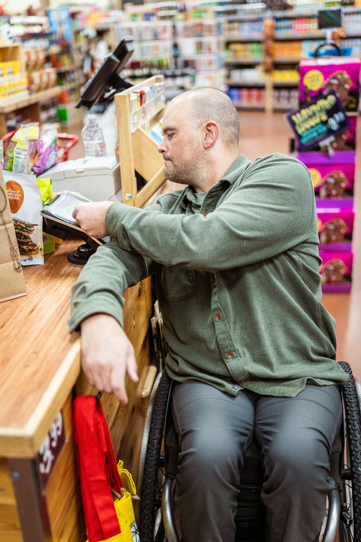 A wheelchair user in a green jacket is standing at a checkout counter in a grocery store, focused on a task at hand.
