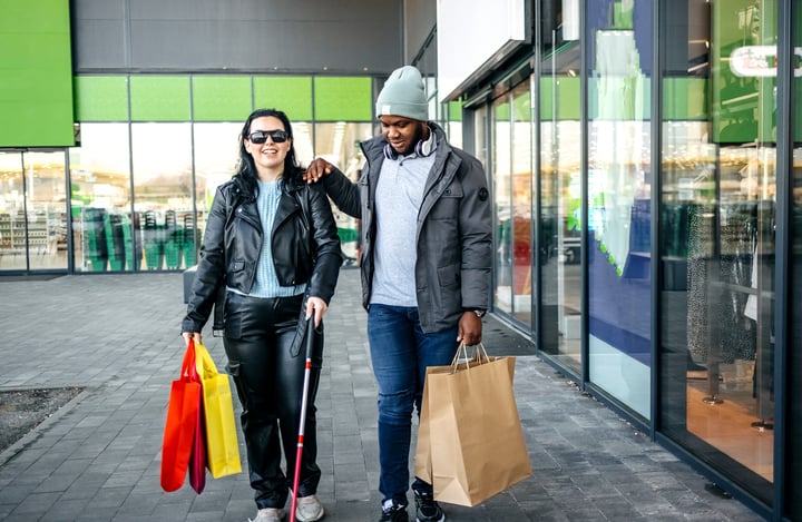 Two people, a man and a woman, are walking together while carrying shopping bags. The woman is wearing sunglasses and holding a cane.