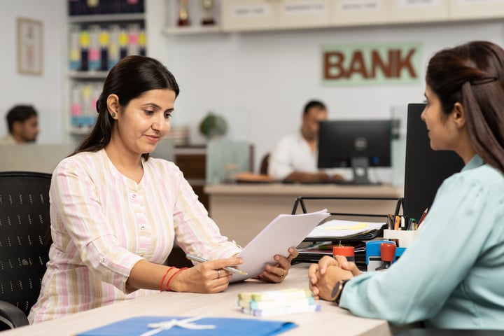 A woman in a pink shirt is sitting at a bank counter, reviewing documents on a tablet. There are other people visible in the background, suggesting this is a banking or financial institution.