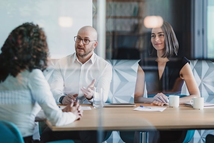 The image shows a group of people having a discussion at a table in a bank. There are two people visible, a man and a woman, both appearing to be engaged in the conversation with a bank teller.