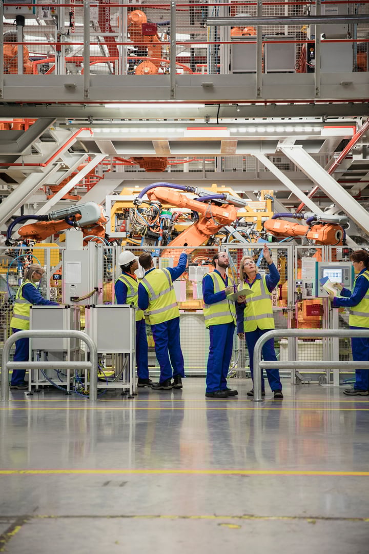 A busy manufacturing facility floor with workers in safety vests operating large robotic machinery and equipment.