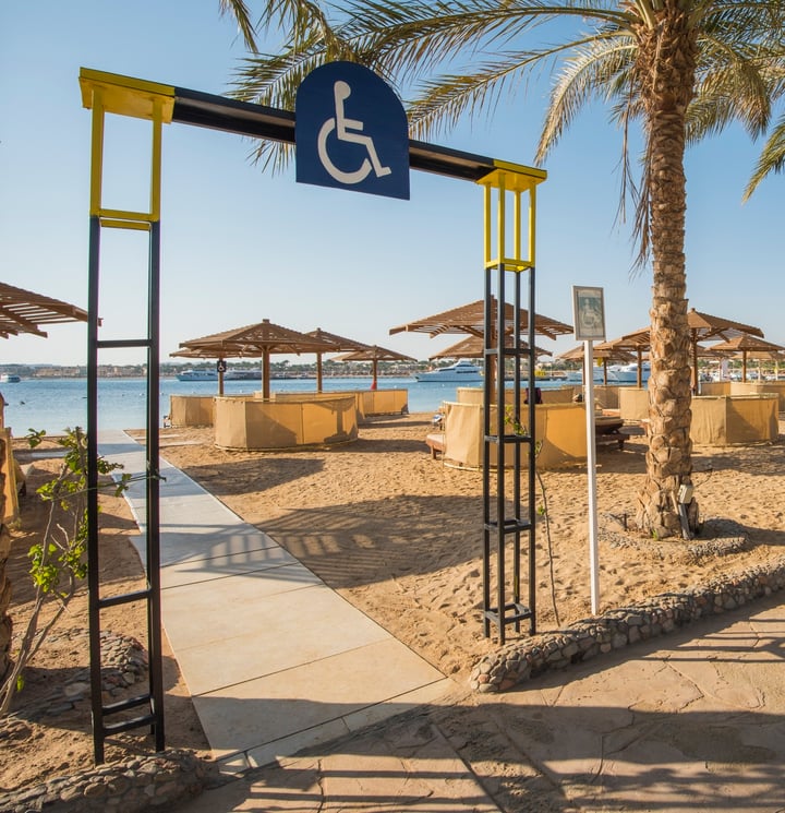 A beach area with palm trees, umbrellas, and a handicap accessibility sign. There is a concrete path along the beach for accessibility.