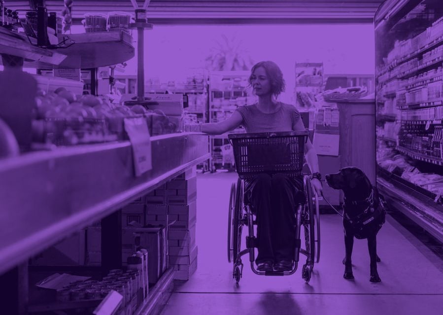 A customer using a wheelchair, accompanied by an assistance dog, shopping in a supermarket aisle
