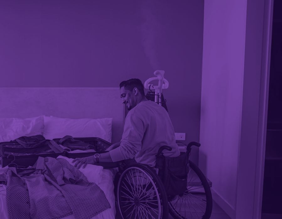 A hotel guest using a wheelchair unpacks a suitcase on the bed in his room