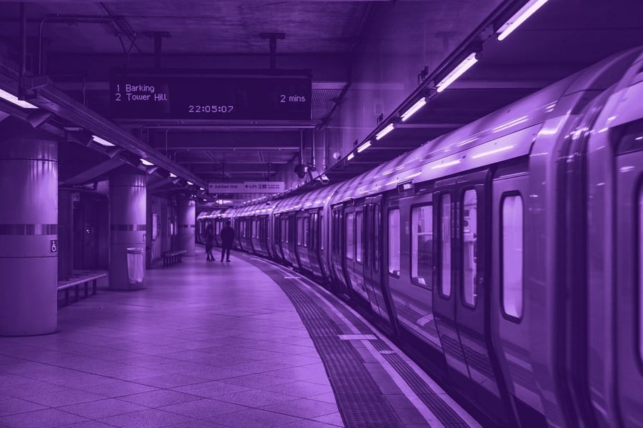 A metro train at an underground platform, next-train board overhead and step-free signage on the pillars