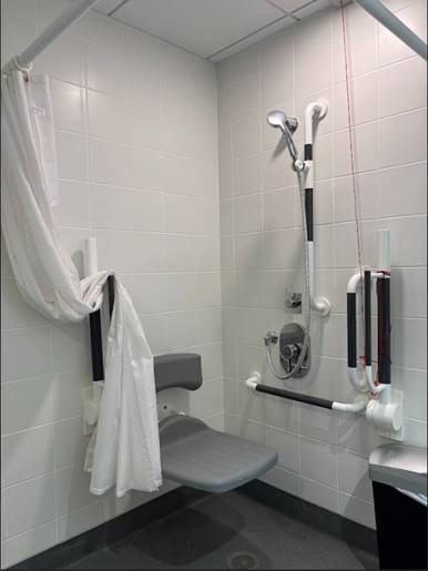 Accessible shower room with a fold-down shower seat, grab bars on the walls, a handheld showerhead, a shower curtain, and a pull cord for assistance.