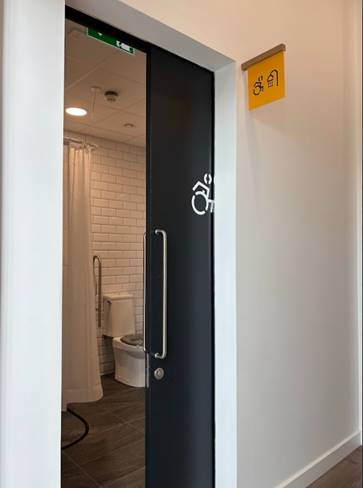 An accessible bathroom with a sliding door partially open, showing a toilet, grab bars, and a shower curtain inside. A yellow accessibility sign with a wheelchair symbol is mounted on the wall above the door.