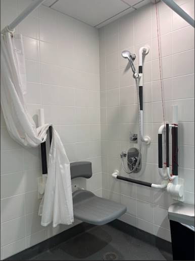An accessible roll-in shower with a fold-down seat, grab bars on the wall, a handheld showerhead, and an emergency pull cord. A shower curtain is tied back to the side.