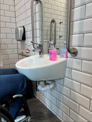 A person in a wheelchair using a small wall-mounted sink in an accessible bathroom. Grab bars are mounted on the wall, and soap dispensers are placed on the sink. There is clear knee space below the basin for wheelchair access.