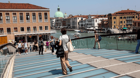 People walking across the modern glass and steel Constitution Bridge (Ponte della Costituzione) over the Grand Canal in Venice, Italy, with historic buildings and boats along the water in the background.