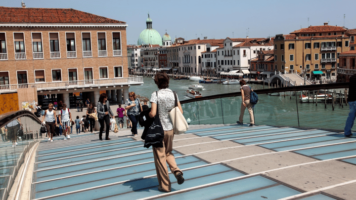 People walking across the modern glass and steel Constitution Bridge (Ponte della Costituzione) over the Grand Canal in Venice, Italy, with historic buildings and boats along the water in the background.