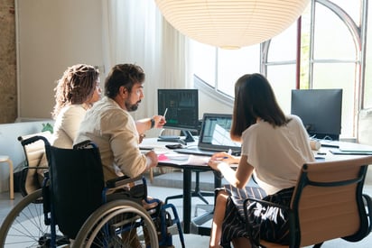 Three colleagues, one a wheelchair user, reviewing data on screens around a meeting table in a bright office